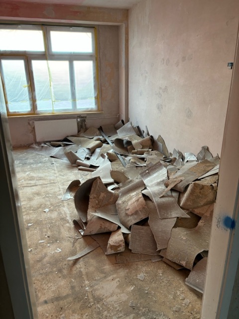Das Bild zeigt ein leergeräumtes Zimmer im Renovierungsprozess. Der Boden ist mit abgetragenen alten Bodenbelag bedeckt. Die Wände sind abgeschliffen. Im Hintergrund ist ein großes Fenster zu sehen, das mit Folie abgedeckt ist, um es vor Staub oder Farbe zu schützen. Der Raum wirkt etwas staubig, und das natürliche Licht, das durch das Fenster hereinfällt, beleuchtet die Szene. Es handelt sich offensichtlich um eine Baustelle im Inneren eines Gebäudes.