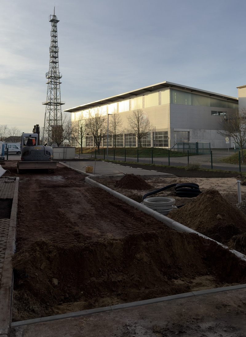 Das Bild zeigt im Vordergrund eine Baustelle, auf der ein neuer Parkplatz entsteht. Der Boden ist teilweise aufgerissen und mit braunem Erdreich bedeckt. Links auf dem Bild sieht man einen Radlader der für die Erdarbeiten genutzt wird. Auf der Baustelle sind helle, längliche Grenzsteine zu sehen, die den neuen Weg und die Parkflächen abgrenzen sollen. Erste Parkplätze sind bereits fertiggestellt und erkennbar. Im Hintergrund befindet sich eine große Übungshalle mit grauer Fassade und vielen Fenstern. Vor der Halle stehen einige kahle Bäume, was auf Herbst oder Winter hinweist. Links im hintergrund ragt ein hoher Metallgittermast in den Himmel, vermutlich ein Funk- oder Sendemast. Der Himmel ist klar und leicht bläulich, die Sonne scheint von links und wirft lange Schatten.