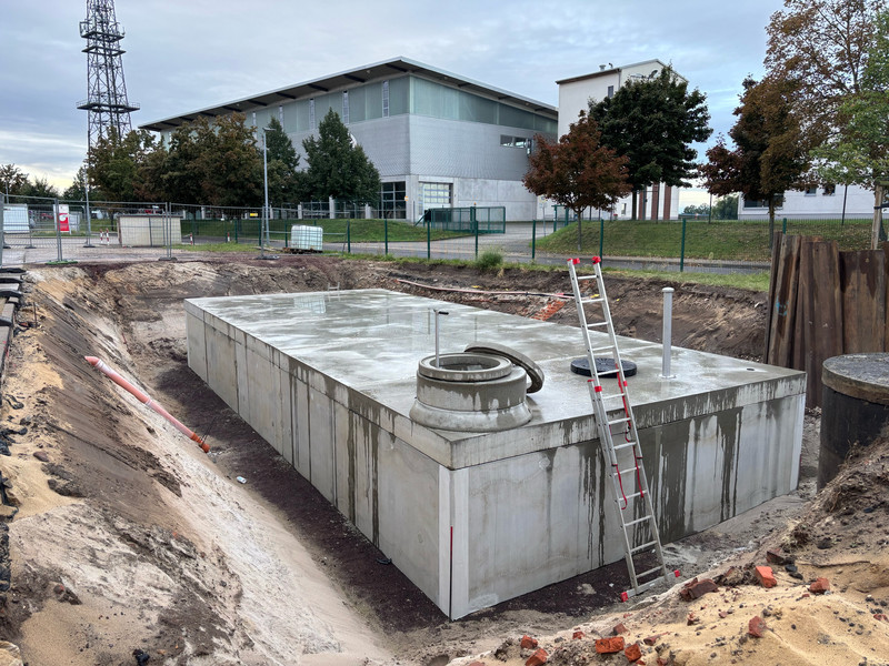 große Baugrube mit Zisterne aus Beton. An die Zsiterne ist eine Leiter angelehnt. Im Hintergrund ist die Übungshalle des IBK zu sehen.