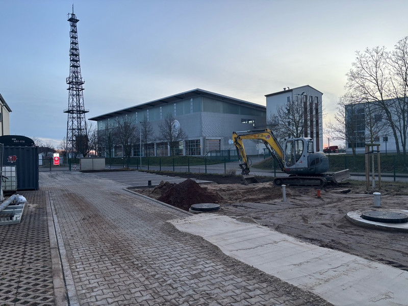 Ein Bagger arbeitet an neben neu gepflasterten Flächen. Eine Übungshalle und ein Funkmast sind im Hintergrund. 