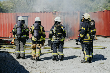 4 Feuerwehrangehörige, ausgerüstet mit schwerem Atemschutz, stehen vor einem rauchenden Container