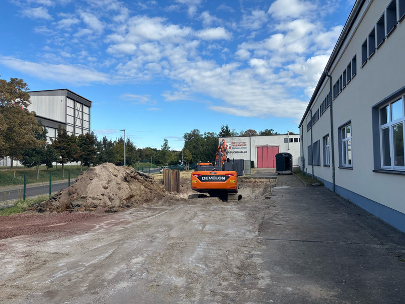 Ein oranger Bagger befindet sich in einem gegrabenen Loch nah an einem Gebäude. Man sieht den Bagger von hinten. Im Hintergrund sind eine Straße und Gebäude des IBK.