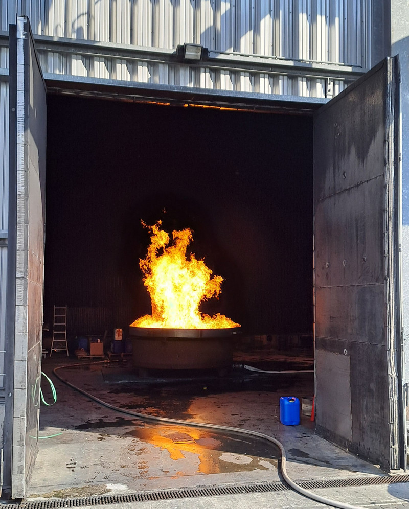 Es steht eine runde Brandwanne mit ca. 2 Metern Durchmesser in einem Brandraum. In der Randwanne brennt eine Flüssigkeit.