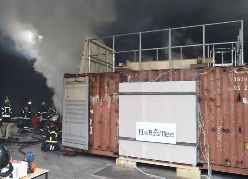 Brandversuch unter Verwendung eines Wand- und eines Deckenbauteils. Das Bild zeigt einen Überseecontainer an dem eine Holzwand angebaut ist, die von einem Feuer im Inneren befeuert wird.