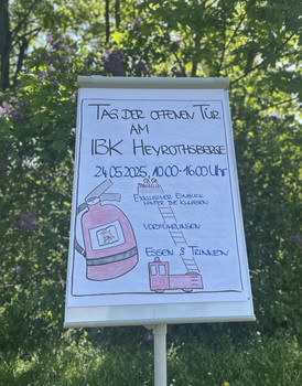 Ein Plakat auf einem Aufsteller, der eine Drehleiter und einen Feuerlöscher zeigt. Die Überschrift sagt Tag der offenen Tür am IBK Heyrothsberge. Dieser ist am 24.04.2025 von 10 bis 16 Uhr.