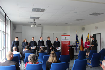 Direktor verliest Schriftstück vor Anwärtern, Ministerin und Publikum.