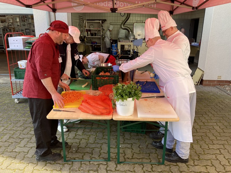 4 Personen stehen in Kochoutfits an einem Tisch und schneiden Gemüse. Im Hintergrund ist eine Feldküche in einer Garage zu sehen.