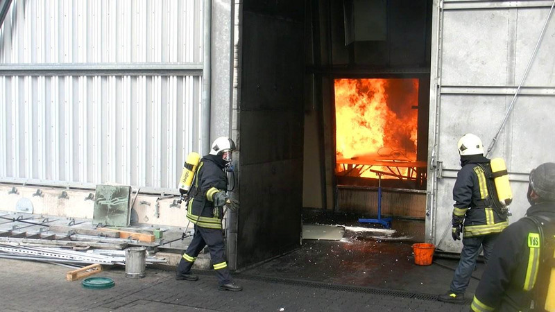 Ein Feuer im Brandhaus wird durch zwei Mitarbeiter vor der Tür des Hauses beobachtet. 