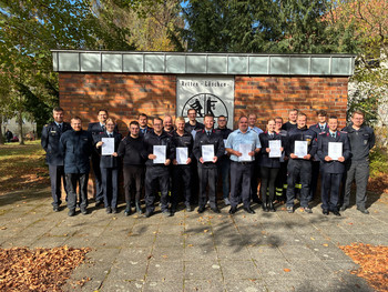 Gruppe von Feuerwehrleuten in Uniform steht vor einer Backsteinwand mit einem Schild, das die Worte 'Retten - Löschen' und ein Feuerwehrsymbol zeigt.