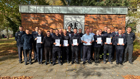 Gruppe von Feuerwehrleuten in Uniform steht vor einer Backsteinwand mit einem Schild, das die Worte 'Retten - Löschen' und ein Feuerwehrsymbol zeigt.