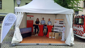 Stand des IBK in einem Zelt mit 3 Mitarbeitern, 2 Stehtischen und Ausstellungsstücken